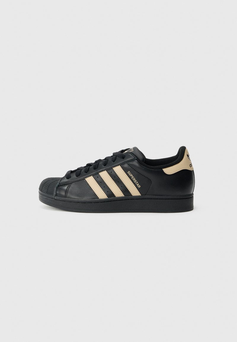 Černá teniska Adidas Superstar s béžovými pruhy, designem špičky s vnější ochranou a značkovým poutkem na patě na bílém pozadí, boční pohled.