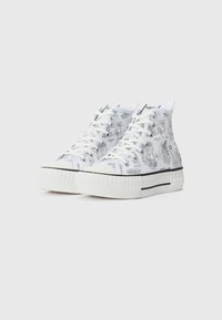 Hvite høyhælte sneakers med trykte svarte designs, med en tykk teksturert såle og hvite snørebånd. Svart aksenter langs sålekanten.