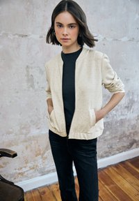 Cazadora bomber cremosa con textura moteada, puños acanalados y dos bolsillos laterales, llevada sobre un suéter negro acanalado y vaqueros oscuros.