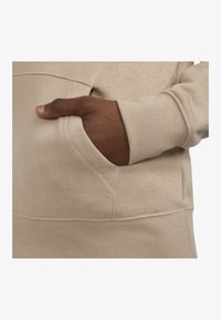 Felpa con cappuccio beige in tessuto morbido, con tasca a marsupio e una mano appoggiata all'interno. Cuciture visibili e polsini a costine.