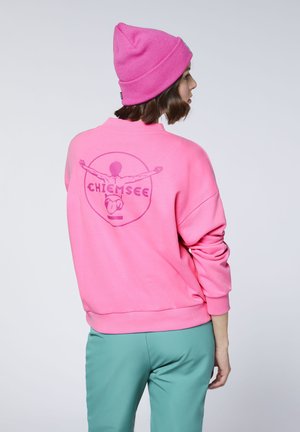 Person facing away wearing a pink beanie and sweatshirt with a graphic of a figure and text on the back, paired with teal pants.