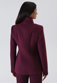 Giacca aderente bordeaux con colletto alto e cuciture strutturate, realizzata in un tessuto liscio. La parte posteriore presenta linee eleganti e pulite.