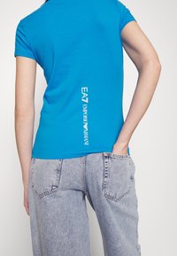 Modré bavlněné tričko s přiléhavým střihem a krátkými rukávy, s bílým textovým logem na straně. Nosit s světle modrými džínami.