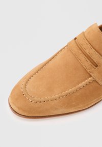 Ταν δερμάτινο μοκασίνι με σχεδιασμό δακτύλου μοκασίνου και ραφές σε απλό λευκό φόντο.