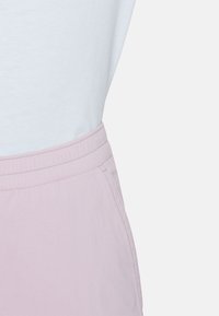 Κοντινή λήψη παντελονιού με ελαστική μέση σε ανοιχτό ροζ, φορεμένο με λευκό πουκάμισο μπουκλωμένο μέσα, που δείχνει πλαϊνή τσέπη και λεπτομέρειες ραφής.