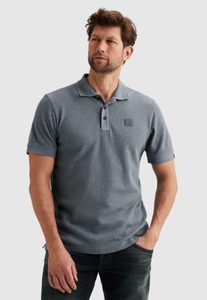 Mann mit Bart, der ein strukturiertes graues kurzärmeliges Poloshirt und dunkle Jeans trägt, mit einer Hand in der Tasche vor einem schlichten Hintergrund stehend.