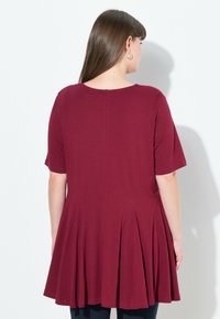 Burgundská tunika s krátkými rukávy, nabíranými záhyby u spodního okraje, kulatým výstřihem a měkkou látkou. Čistý design zadní části s detaily švu.