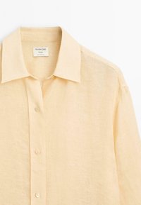 Helles beigefarbenes Leinenhemd mit spitzem Kragen, Knopfverschluss und leicht transparenter Textur. Etikett im Kragen sichtbar.