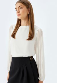 Blusa bianca con maniche a sbuffo lunghe e trasparenti. Tessuto liscio, scollatura rotonda. Abbinata con una gonna nera a vita alta con accenti dorati.