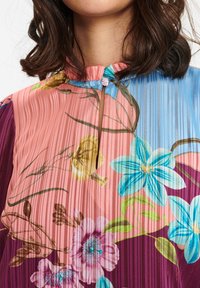 Pleated blouse dengan cetakan bunga berwarna-warni. Memiliki leher tinggi dengan penutupan kancing dan tekstur bergaris dalam warna merah muda, biru, dan ungu.