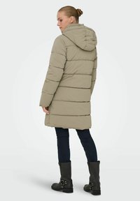 Pufferjakke i lys khaki med vandret syning, hætte og lange ærmer. Båret med mørke jeans og sorte ankelstøvler.