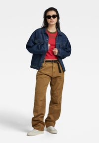 Jeansjack, rode shirt met groene print, kaki broek met zakken, witte sneakers en zwarte zonnebril, tegen een effen achtergrond.