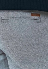 Szary teksturowany materiał z subtelny wzorem, z kieszenią i brązową skórzaną metką marki. Ciemny prążkowany materiał powyżej linii talii.