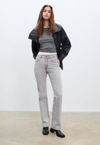 Crna kožna jakna, siva skraćena majica, svijetlosiva ravna traperice, crne čizme do gležnja, na običnoj bijeloj pozadini.