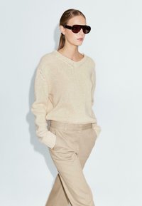 Beige gebreide trui met een V-hals, gecombineerd met lichtbeige broeken. Model draagt grote, donkere zonnebril. Schone, minimalistische achtergrond.