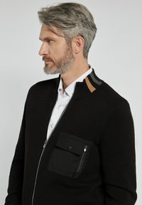 Czarna kurtka zapinana na zamek z teksturowanej tkaniny, kontrastującymi beżowymi i szarymi akcentami na kołnierzu oraz przednia kieszeń zapinana na nap oraz zamek.