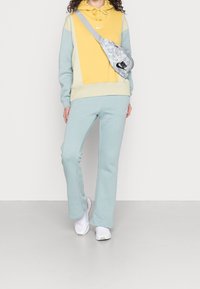 Pantaloni della tuta azzurri e una felpa con cappuccio gialla, grigia e color crema con coulisse. Un marsupio grigio mimetico con un logo nero, indossato a tracolla.