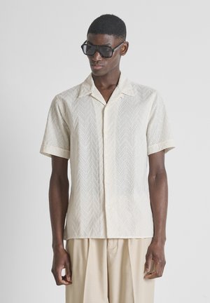 Jonge man met zwarte zonnebril, crèmekleurig overhemd met korte mouwen en chevron oogjespatroon, en beige broek tegen een effen achtergrond.