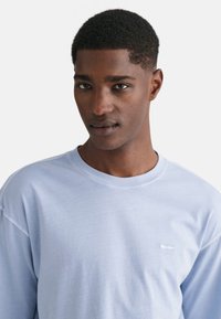 Světle modré tričko s dlouhým rukávem z měkké bavlny, s kulatým výstřihem a jemným logem na hrudi. Pohodlný střih se švy.