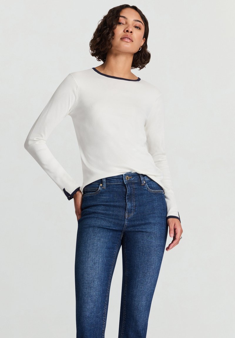 Langärmliges weißes Oberteil mit navyfarbenem Ausschnitt und Bündchen, kombiniert mit dunkelblauen Jeans. Glatter Stoff mit figurbetontem Schnitt.