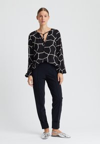 Zwarte blouse met wit geometrisch patroon, lange mouwen, strik halslijn; gecombineerd met een aansluitende zwarte broek en zilveren metallic platte schoenen.