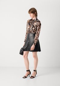 Blusa a maniche lunghe in stampa zebra marrone e beige con colletto, abbinata a una gonna a campana in pelle nera, sandali con tacco alto neri e una borsa piccola.