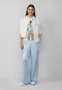 Veste en denim blanc sur un t-shirt graphique, pantalon ample en satin bleu clair, et baskets blanches, créant un look décontracté en couches.