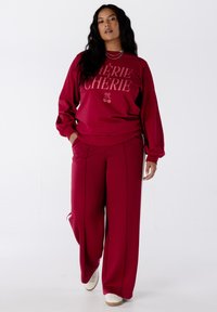 Femme marchant dans un sweat-shirt rouge avec le texte "CHÉRIE" et des cerises, portant un pantalon large assorti, des baskets blanches et des colliers en argent superposés.