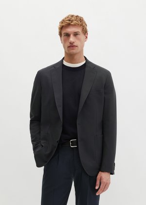 Mann mit kurzen blonden Haaren, der einen dunklen Blazer über einem schwarzen Pullover und Hose trägt, vor einem schlichten weißen Hintergrund stehend.