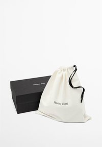 Zwarte rechthoekige doos en witte zak met trekkoord met zwarte koorden, beide met het label Massimo Dutti, op een effen witte achtergrond.