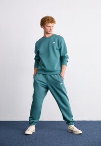 Teal sweatshirt en bijpassende joggers van zacht materiaal. Heeft ribgebreide boorden en een geborduurd logo op de borst en het been.
