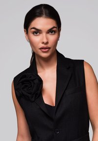 Zwarte mouwloze blazer met een grote stoffen rozendetail op de schouder, voorzien van een geknipt kraag en een sluiting met één knop.