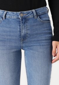 Light blue denim jeans feature a mid-rise waist, front pockets, and subtle fading in the fabric. The design includes a metal button closure.