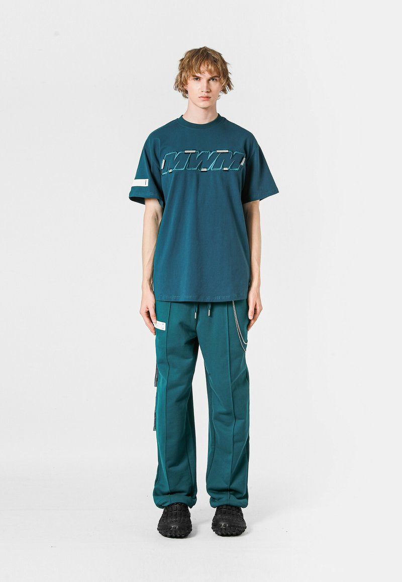 Camiseta de manga corta color verde azulado con un gráfico blanco audaz, combinada con pantalones de color verde azulado a juego que presentan bolsillos y un detalle de cadena plateada. Zapatos negros.