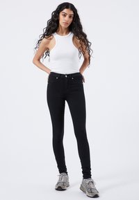 Jean skinny noirs taille haute, à cinq poches, avec des accessoires en métal argenté, associés à un débardeur blanc à côtes et des baskets grises.