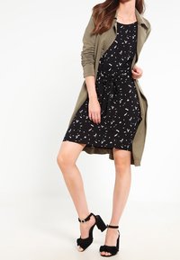 Robe noire avec motif abstrait blanc, taille cintrée, associée à un long cardigan vert olive et des sandales à talons hauts noires.