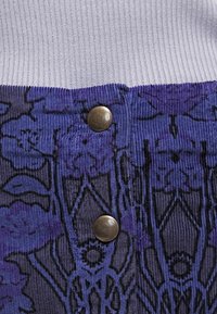 Långhårig klädesplagg i lila med ett blommönster, med en vit ribbad övre kant och två runda metallknappar fram.