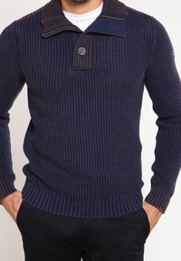 Granatowy sweter wełniany o teksturowanej powierzchni, z kołnierzem na pół-zamek oraz ściągaczami na mankietach i u dołu, z pojedynczym guzikiem na kołnierzu.