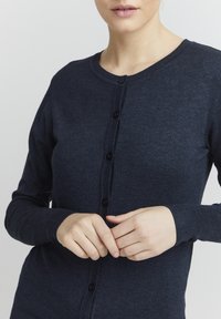 Cardigan bleu marine foncé avec un col rond, fermeture à boutons sur le devant, et des poignets et un ourlet côtelés. Fait en matière tricotée douce.