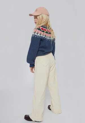 Jersey de punto azul marino con canesú floral, pantalones de pana crema con bolsillos en forma de corazón y gorra rosa. Zapatos negros, bajos de pantalón con flecos.