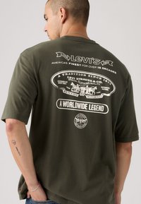 Ελιά πράσινη κοντομάνικη μπλούζα με λευκό γραφικό σχέδιο στην πλάτη, που περιλαμβάνει λεπτομέρειες της μάρκας και εικόνες αλόγων. Απαλή υφή.