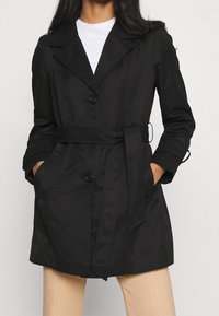 Persona vistiendo un abrigo de trench negro con cinturón sobre una camisa blanca, con las manos en los bolsillos, combinado con pantalones beige frente a un fondo neutro.