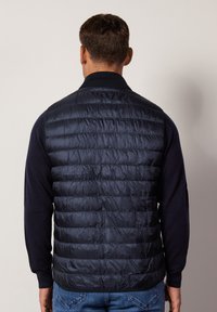 Granatowa kamizelka puchowa o pikowanej fakturze, z wysokim kołnierzem i zapinana na zamek. Noszona na granatowym swetrze, zestawiona z niebieskimi dżinsami.