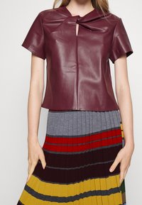 Top in pelle bordeaux con dettaglio a nodo sul collo, maniche corte e una vestibilità rilassata, abbinato a una gonna plissettata multicolore.