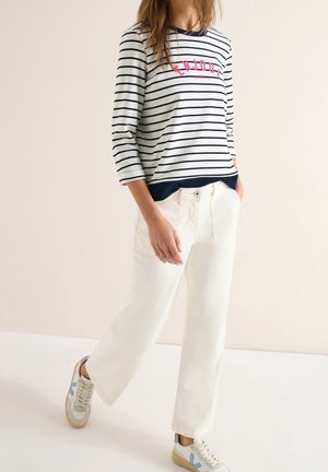 Femme portant un pantalon blanc à jambes larges, une chemise à manches longues rayée avec le texte "ENJOY" et des baskets blanches marchant devant un fond beige uni.
