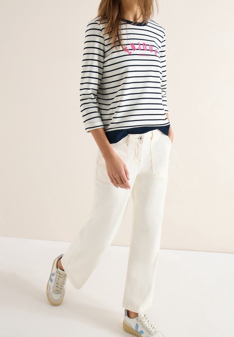 Femme portant un pantalon blanc à jambes larges, une chemise à manches longues rayée avec le texte "ENJOY" et des baskets blanches marchant devant un fond beige uni.