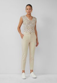 Pantalons slim beige avec une fermeture à bouton, associés à un haut texturé à motifs chevrons beige et or et des baskets blanches.