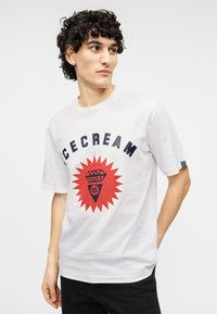 Λευκό βαμβακερό t-shirt με μεγάλο κόκκινο σχέδιο έκρηξης που απεικονίζει διαμάντι και σύμβολο δολαρίου. Μαύρα γράμματα γράφουν "ICECREAM" πάνω από το σχέδιο.