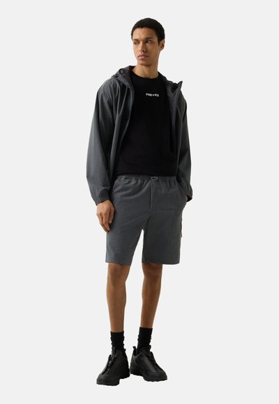 Młody mężczyzna w ciemnoszarej kurtce z kapturem, dopasowanych szortach, czarnych skarpetach, czarnych masywnych butach oraz czarnej koszulce z napisem „FIRE + ICE”.