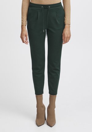 Dark green tapered pants made of soft fabric, featuring a drawstring waist, pleated front, and ribbed cuffs. Brown high-heeled boots complement the look.